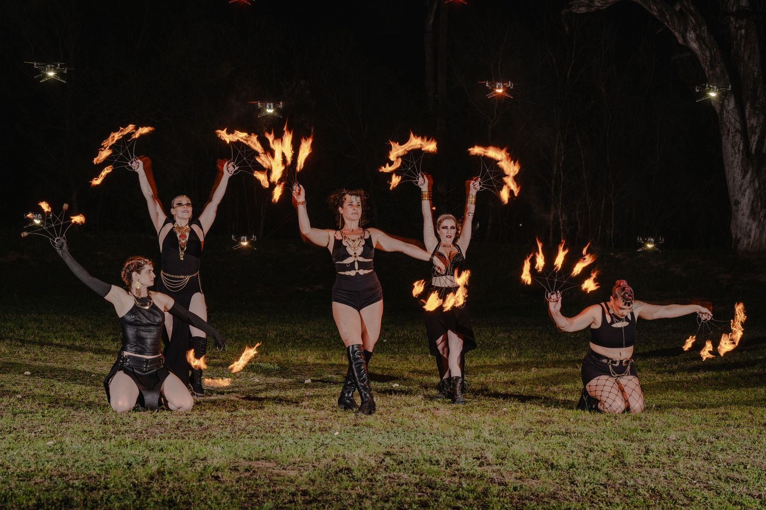 Five female fire dancers perform at night outdoors, spinning flaming props in a dramatic choreographed display.
