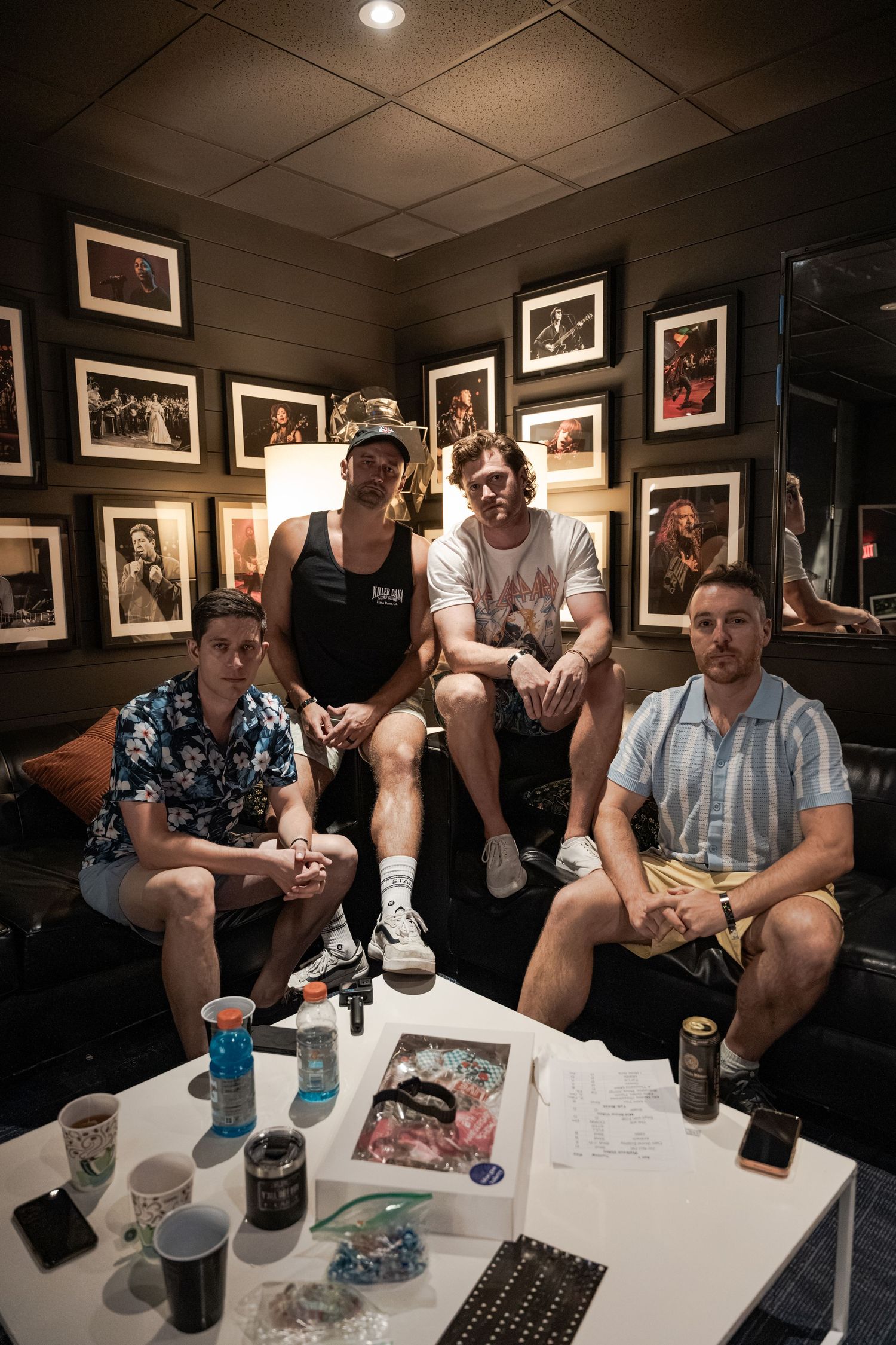 Four young men pose together in a dimly lit room with framed photos on dark walls, drinks on a white table.