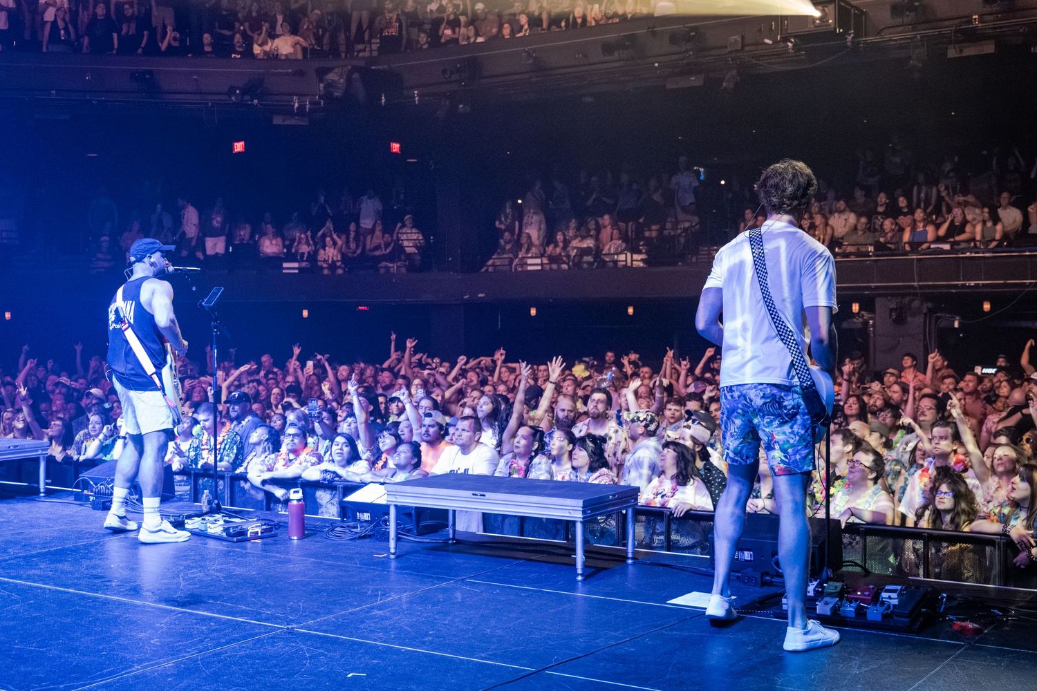 Two performers on stage facing a large energetic crowd at an indoor concert venue with blue lighting.