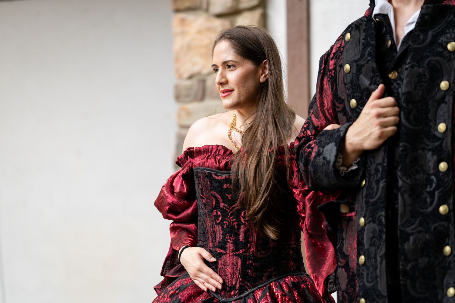 Woman in red and black Victorian corset dress poses with man in ornate velvet costume at outdoor historical event.