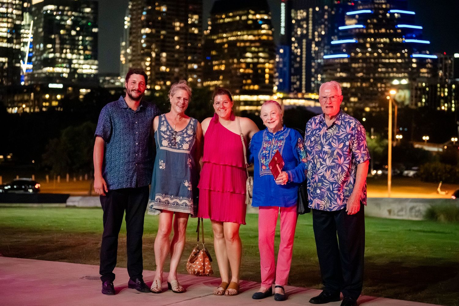 A group of people in colorful attire pose together at night with a city skyline illuminated in the background.