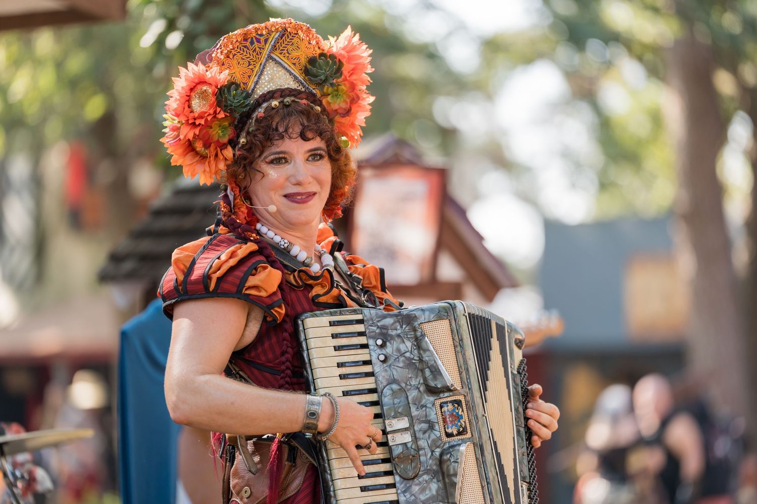 A performer wearing orange flowers plays an accordion at an outdoor festival with trees in the background.