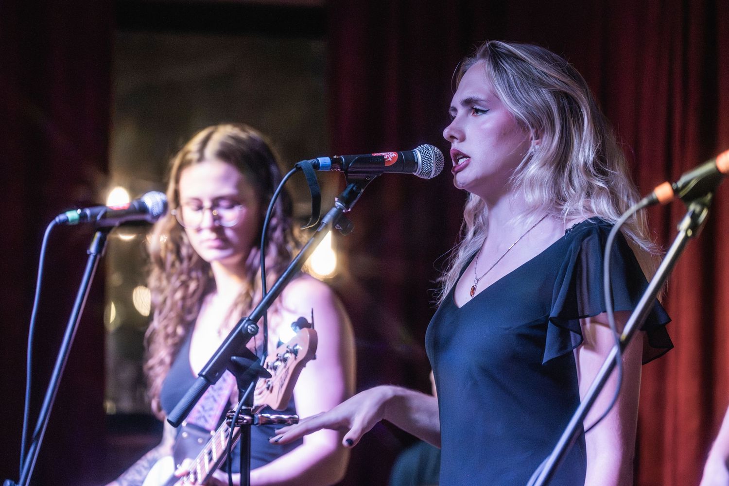 Musicians perform on stage with microphones and instruments at a live music venue with red curtains in the background.
