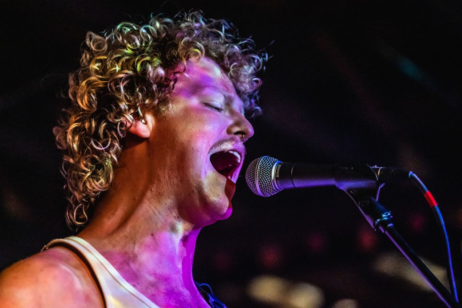 A performer sings passionately on stage with dramatic purple lighting and curly hair illuminated in the spotlight.