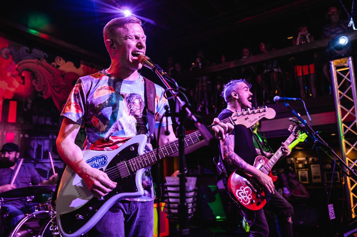 Musicians perform on stage with colorful purple and red lighting during a live concert performance.