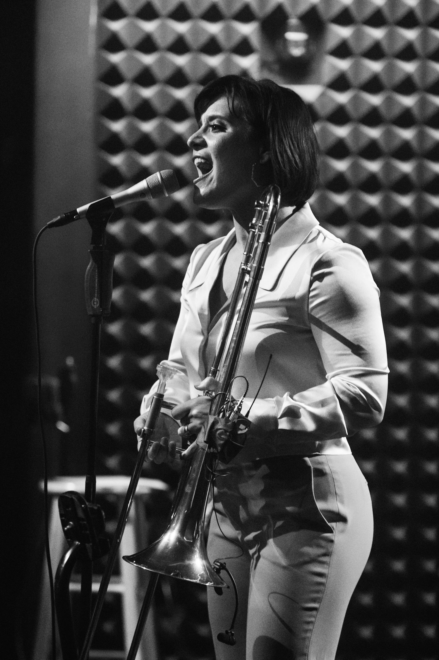 A musician plays trombone while performing in a white outfit against acoustic panels in black and white.