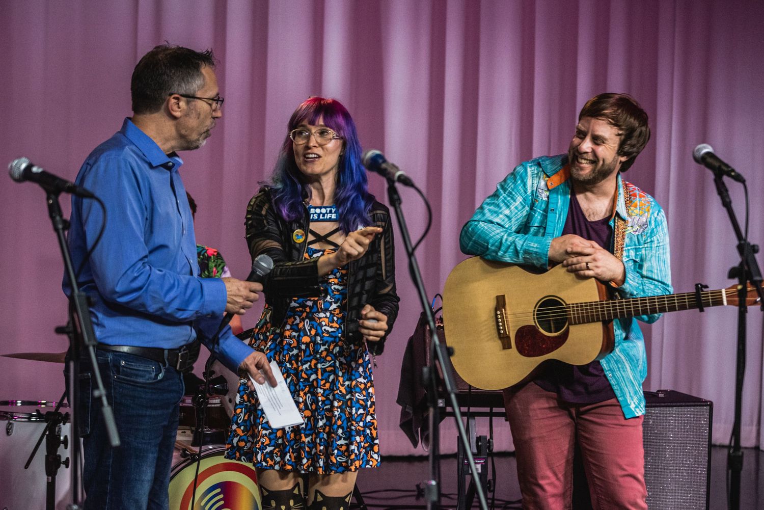 Band members engaged in conversation during performance on stage with pink backdrop.