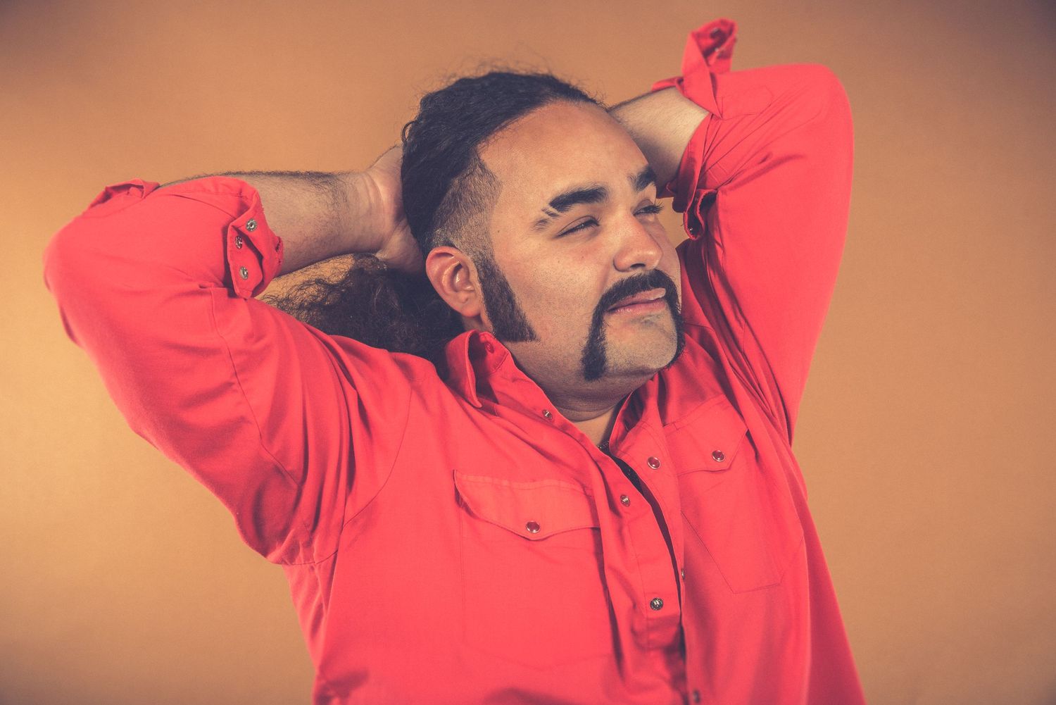 Portrait of someone in a red shirt stretching with hands behind head against orange background.