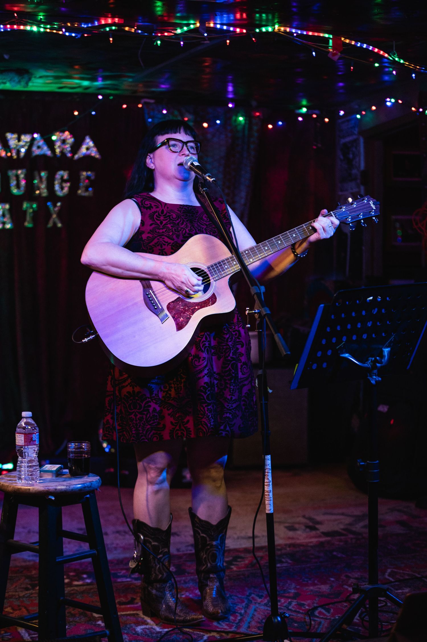 Performer in dress and boots playing guitar on dimly lit stage with string lights.