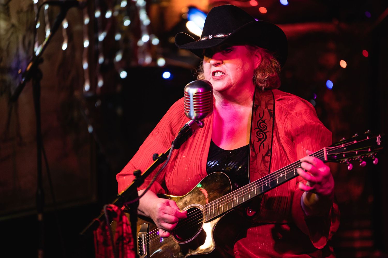 Musician in red jacket and cowboy hat performs on stage with guitar under colorful lighting.