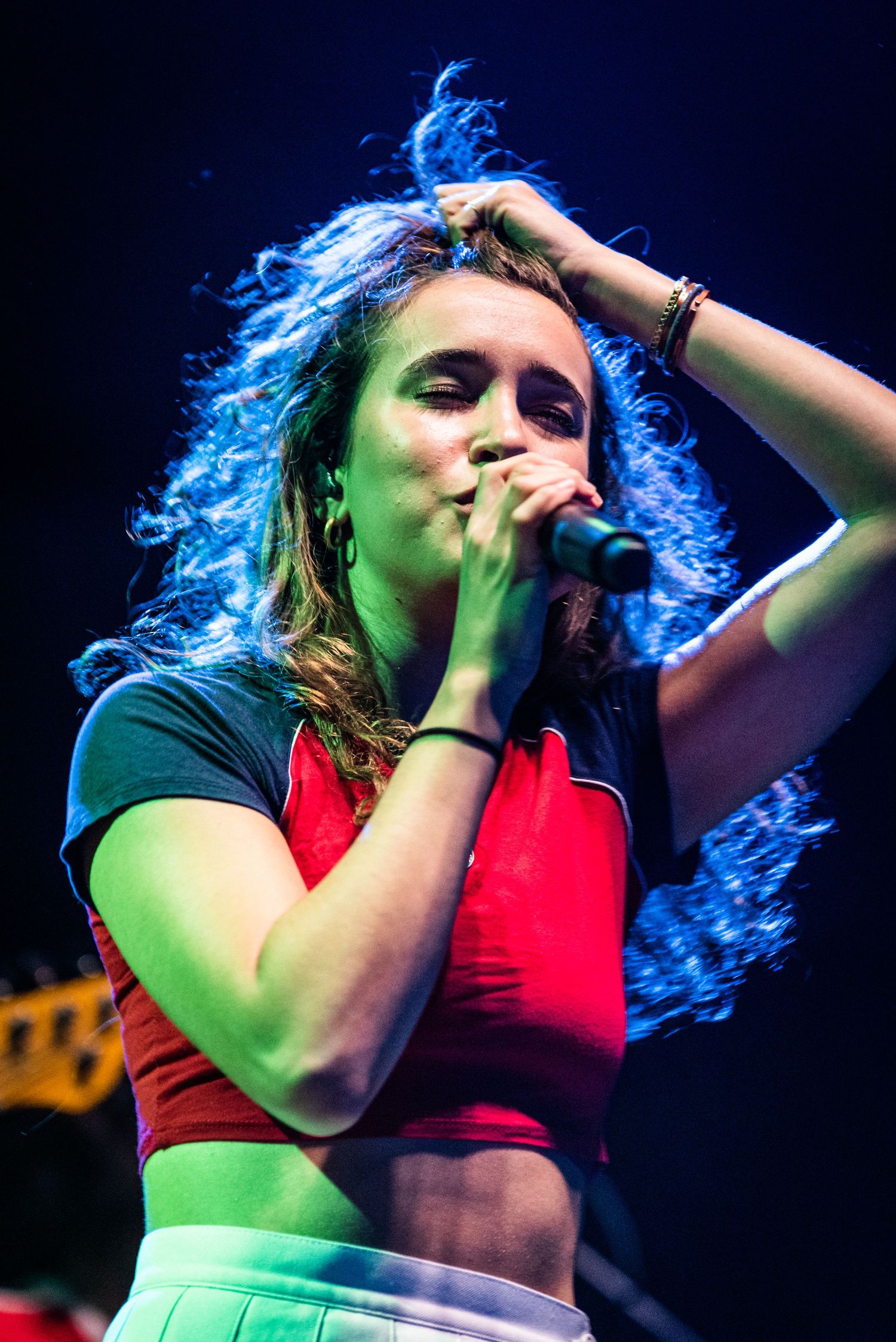 Stage performer with vibrant blue-lit hair sings into microphone wearing crop top during live performance.