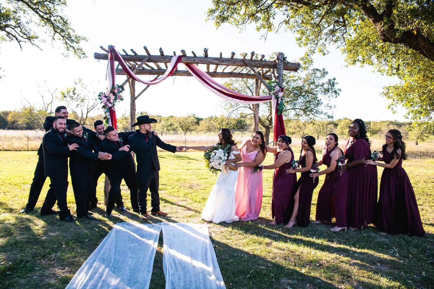 Fun candid wedding party photo with groomsmen and bridesmaids posing playfully under decorated arch.