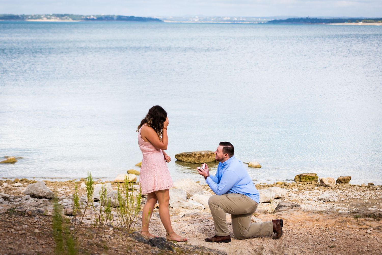A romantic proposal scene captured by the water's edge on a sunny afternoon.