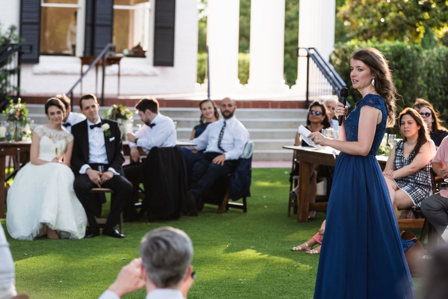 Wedding guest in navy dress gives a speech while couple and guests listen on the lawn.