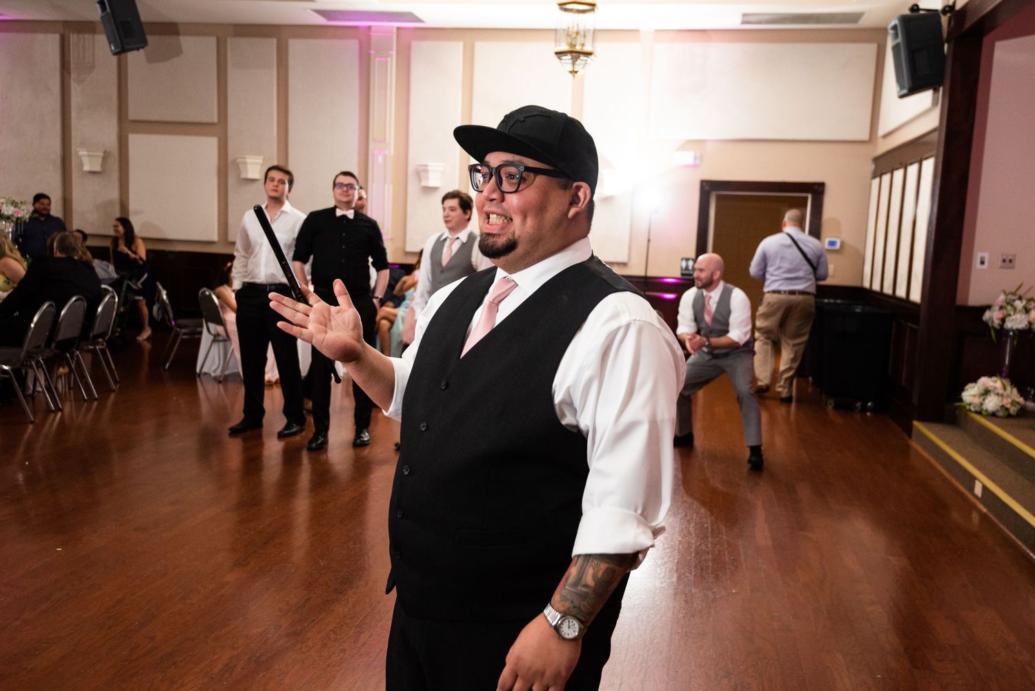 Person in formal attire gestures expressively while standing on wooden dance floor at reception.