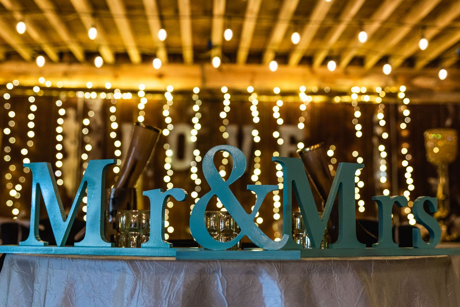 Decorative teal 'Mr & Mrs' letters displayed on a table with string lights in the background.