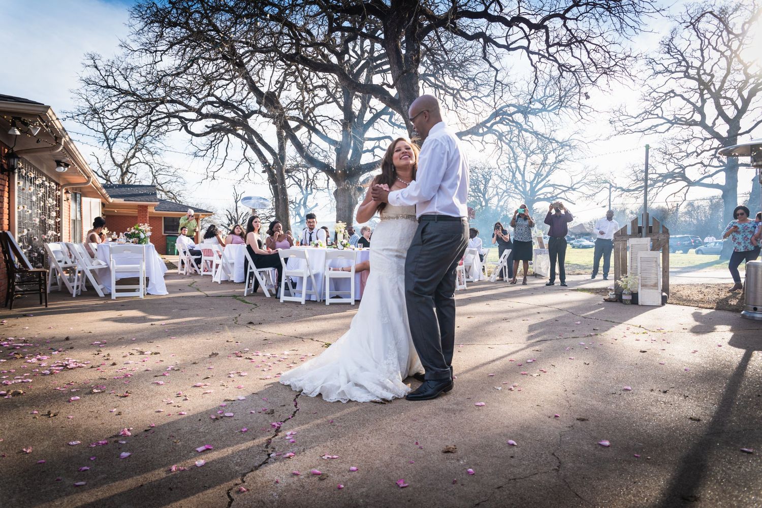 Newlyweds share dance at outdoor reception venue with guest tables and trees in background.