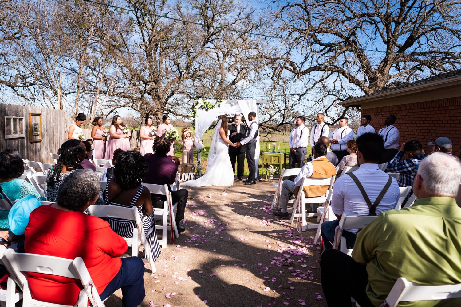 Wedding guests watch the ceremony from white chairs as the couple exchanges vows under sunny skies.