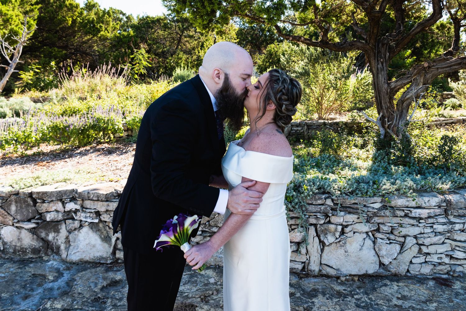 Newlyweds embrace and kiss in a romantic moment outdoors with sunlit garden backdrop.