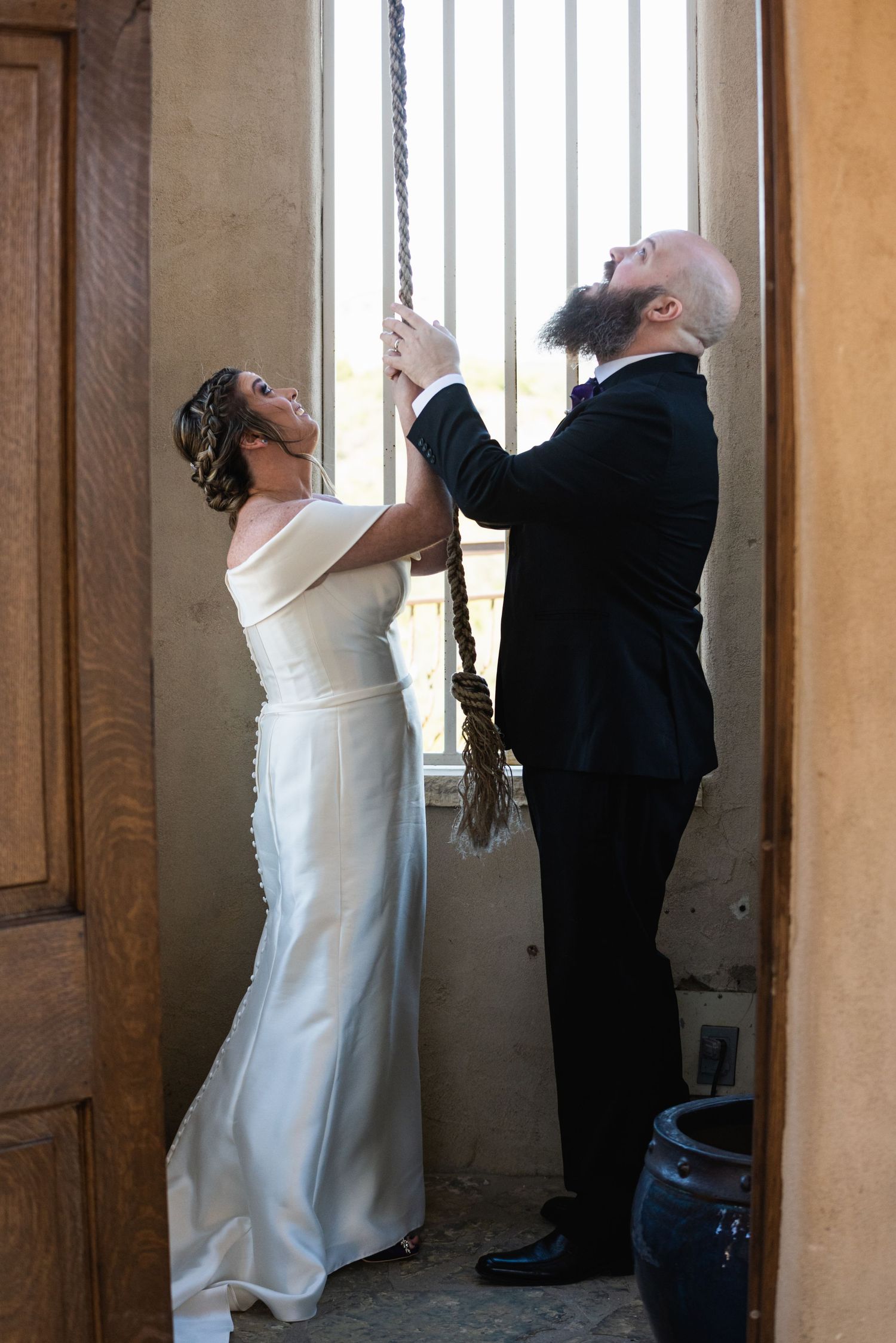 A couple in formal wedding attire rings a bell together in a doorway.