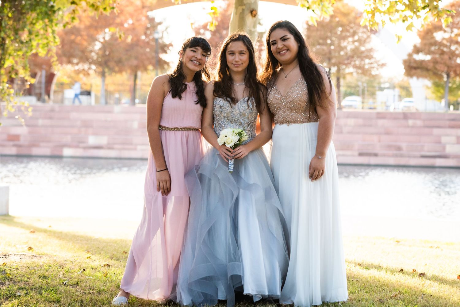Three attendants in pastel formal gowns pose together near water.