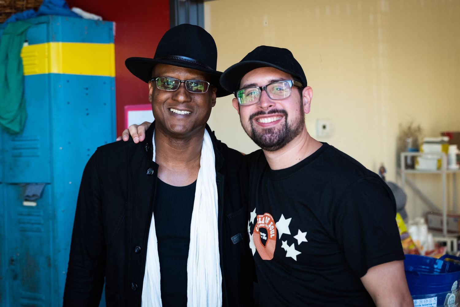 Two musicians posing together backstage wearing black clothing and hats.