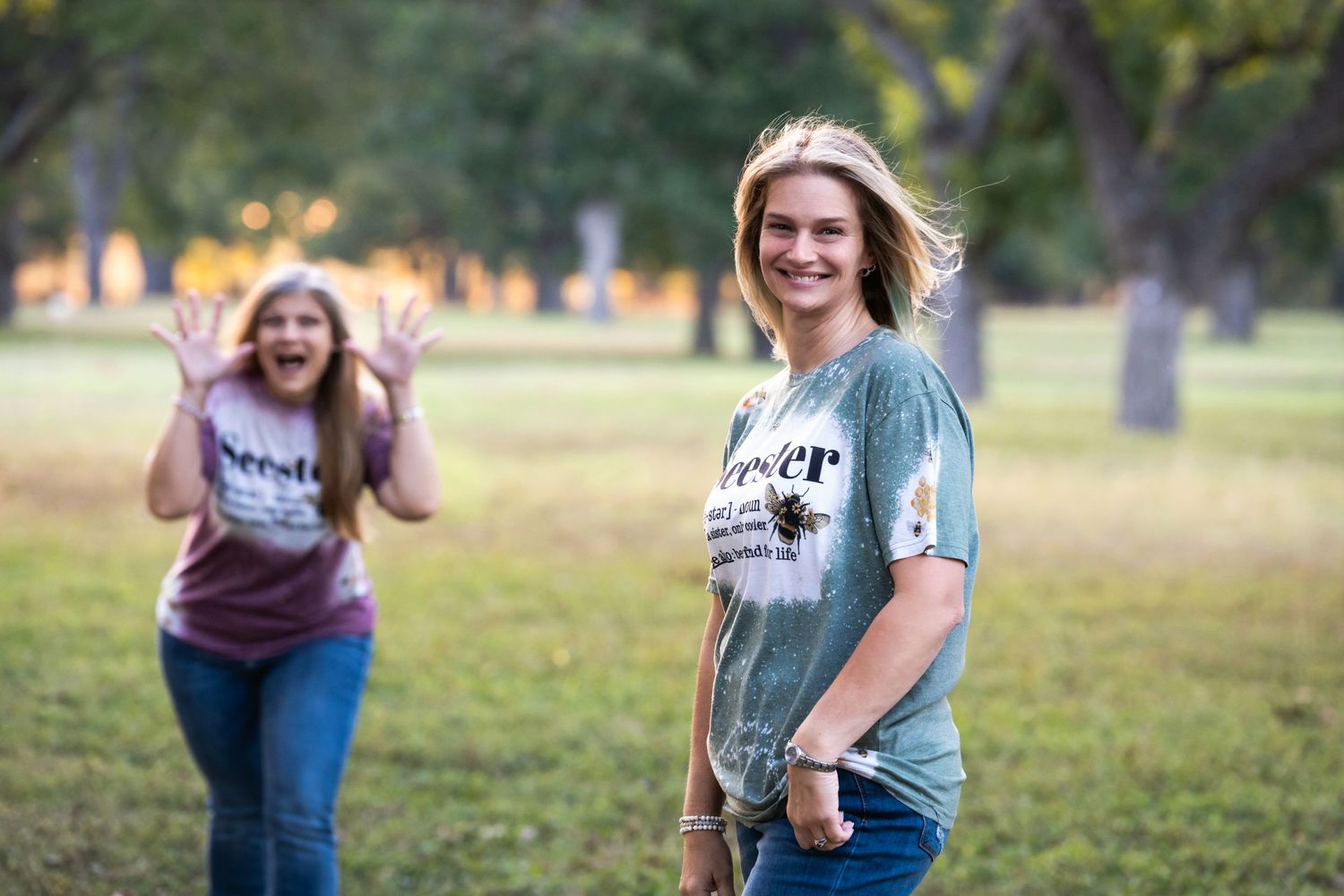 Two people in matching shirts wave and pose while standing in a sunlit park setting.