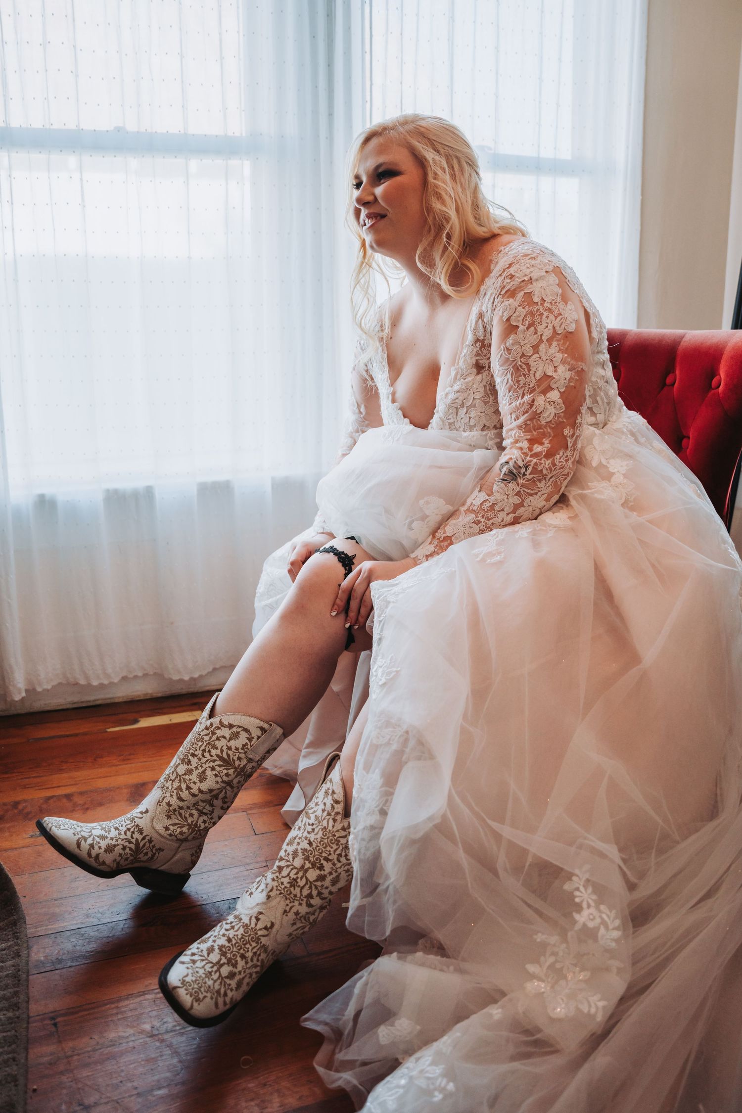 A bride in a lace wedding dress shows off embellished cowboy boots while seated by a window.
