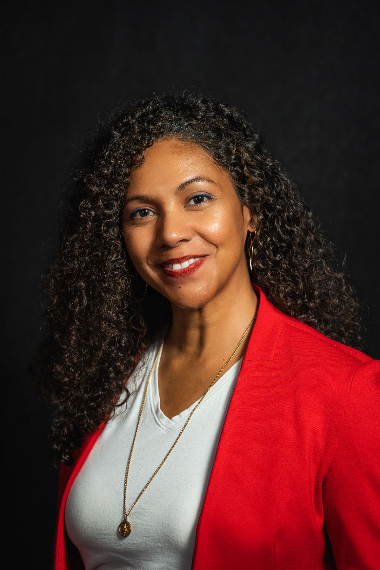 Professional portrait showing person in red blazer and gray top with curly hair against dark background.