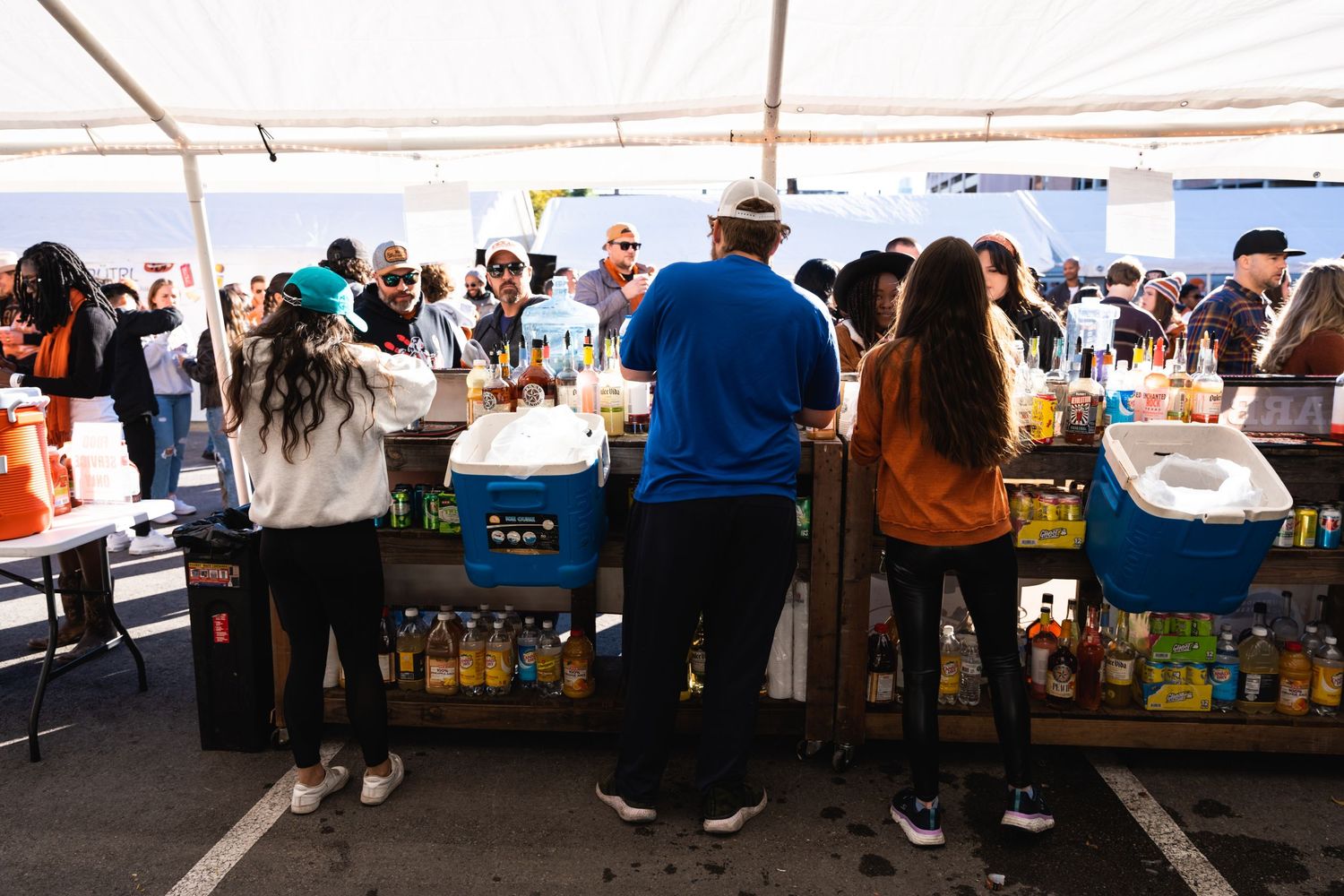 Crowd gathers around vendors serving drinks under a white tent at an outdoor event.