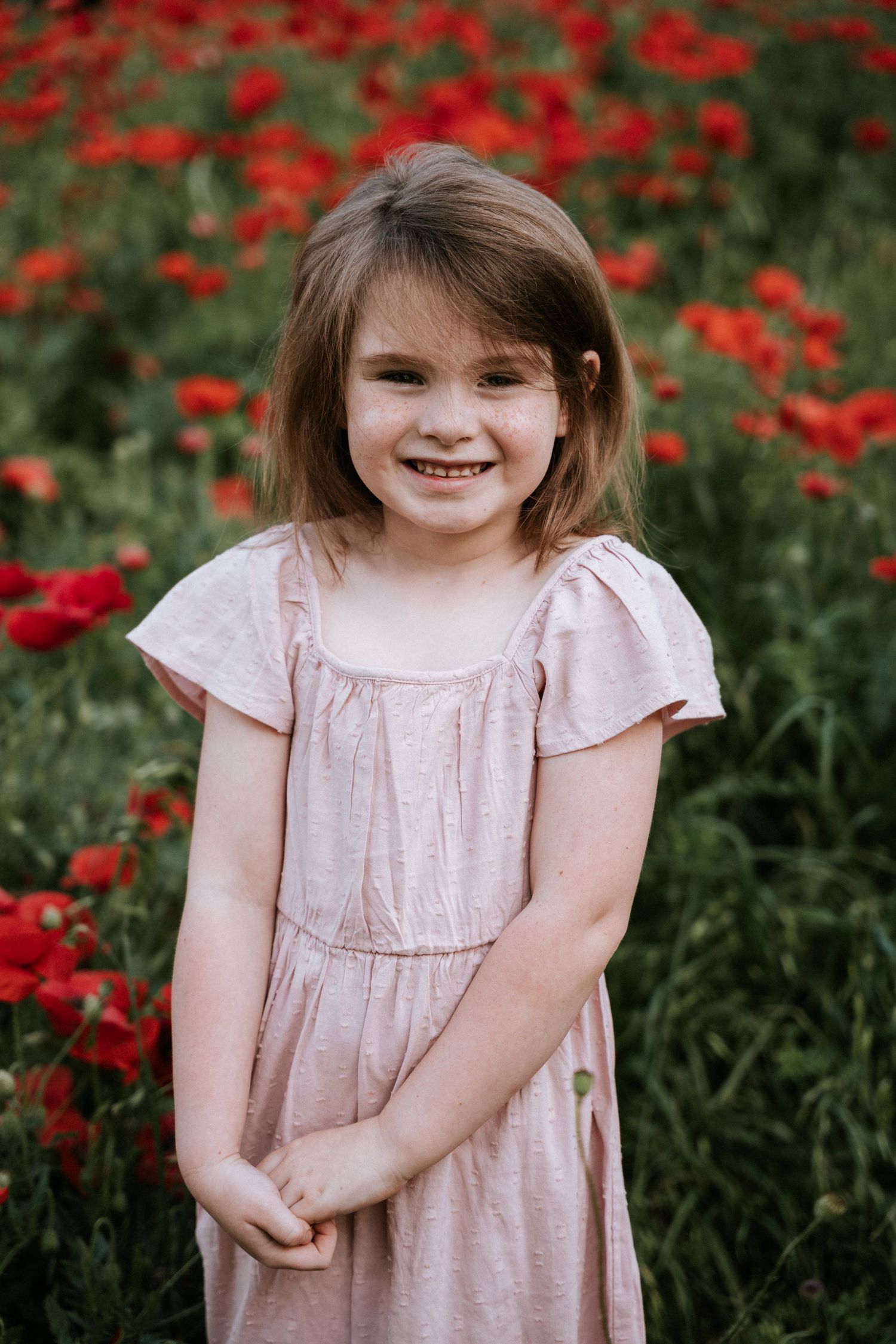 Child in pink dress poses in front of blooming red poppies.