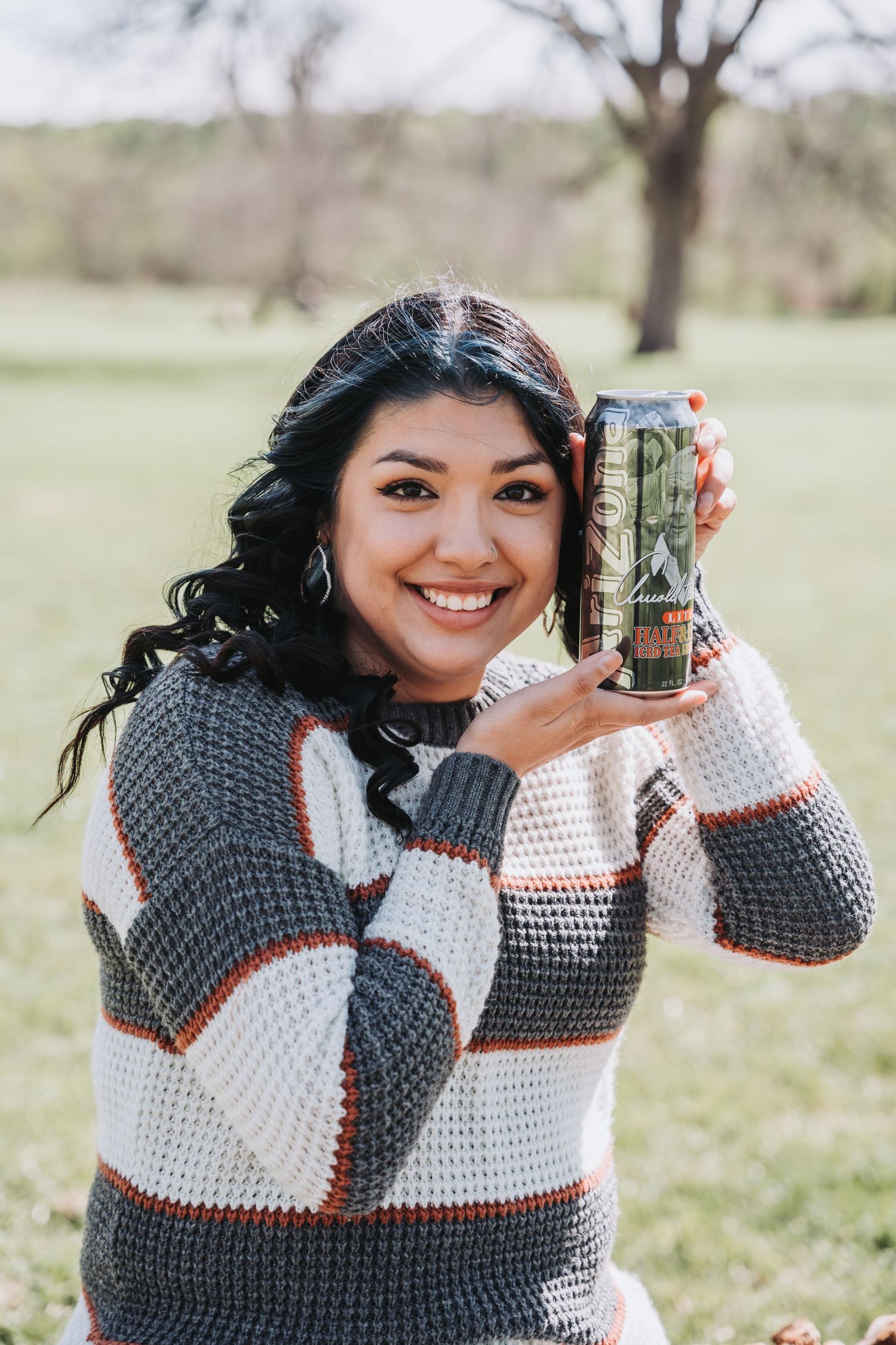 Person in striped sweater holds up a drink while smiling outdoors on sunny day.