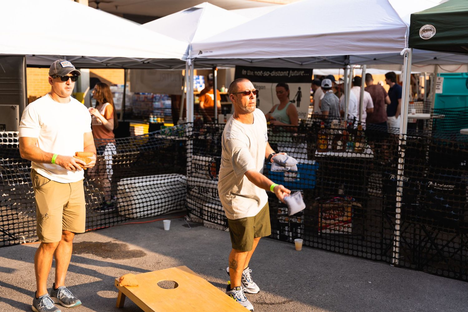 People playing cornhole at an outdoor event with vendor tents and gaming setups.