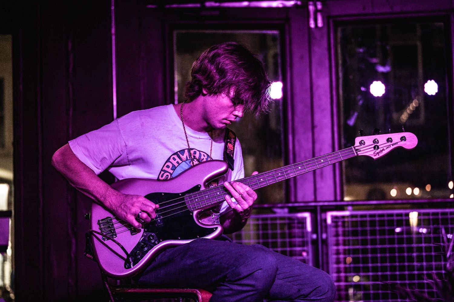 Musician plays bass guitar intensely under purple stage lights in live venue setting.