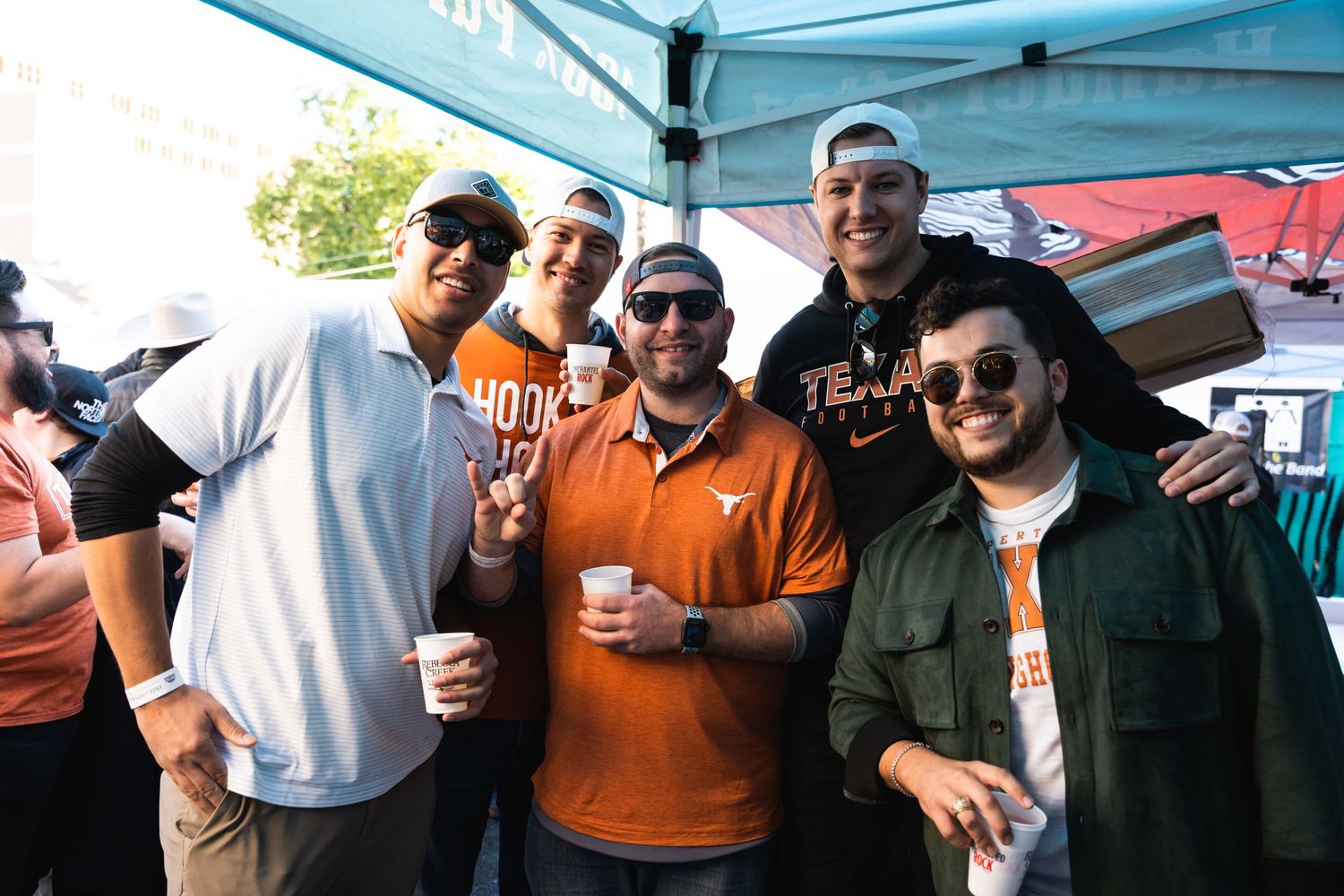 Friends posing together with drinks under a blue tent at an outdoor gathering.