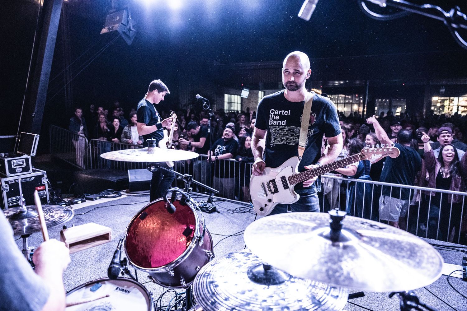 Concert performance with drummer and guitarist on stage surrounded by crowd barriers.