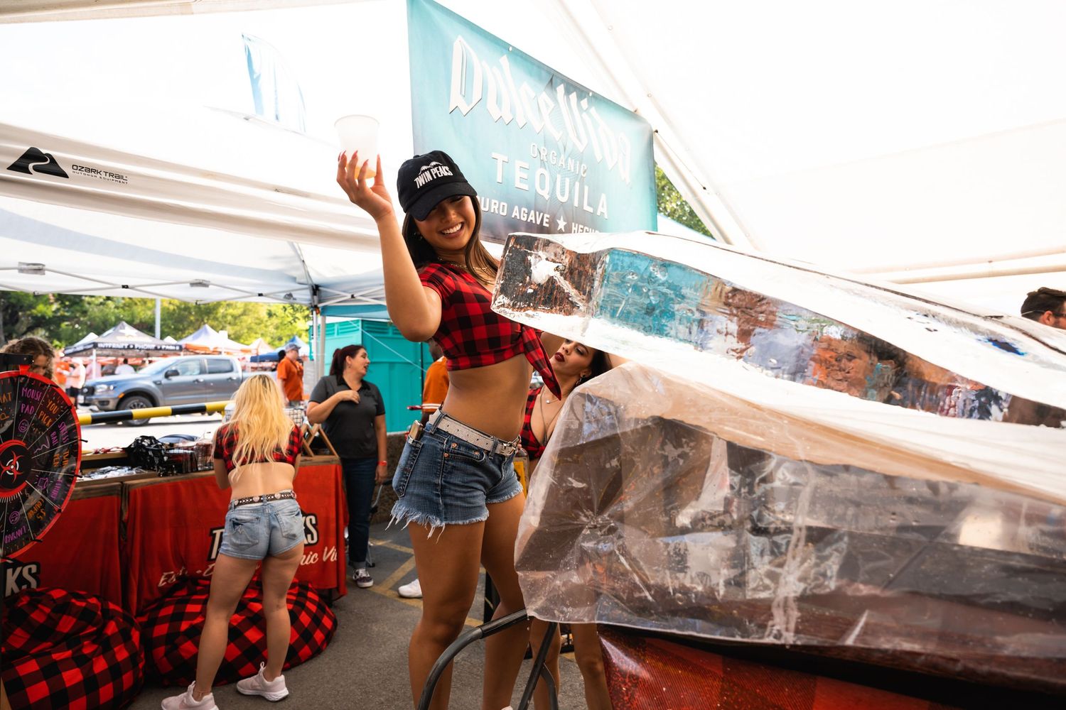 Festival attendee in plaid and denim poses near ice sculpture at outdoor tequila tent.