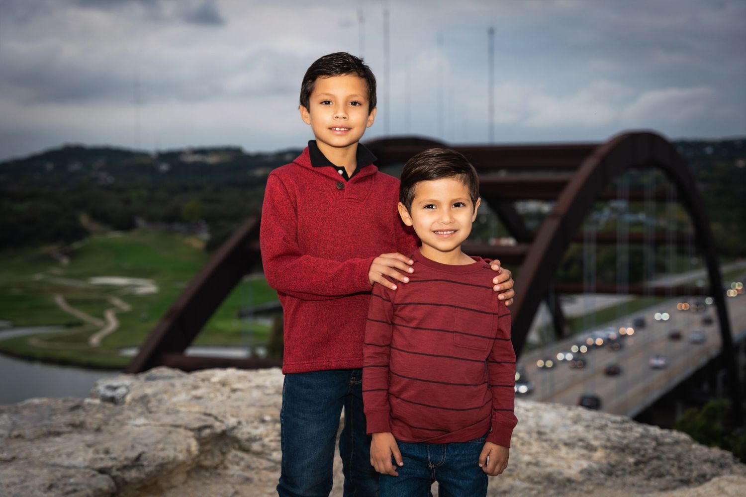 Two people in matching red sweaters pose in front of Pennybacker Bridge.