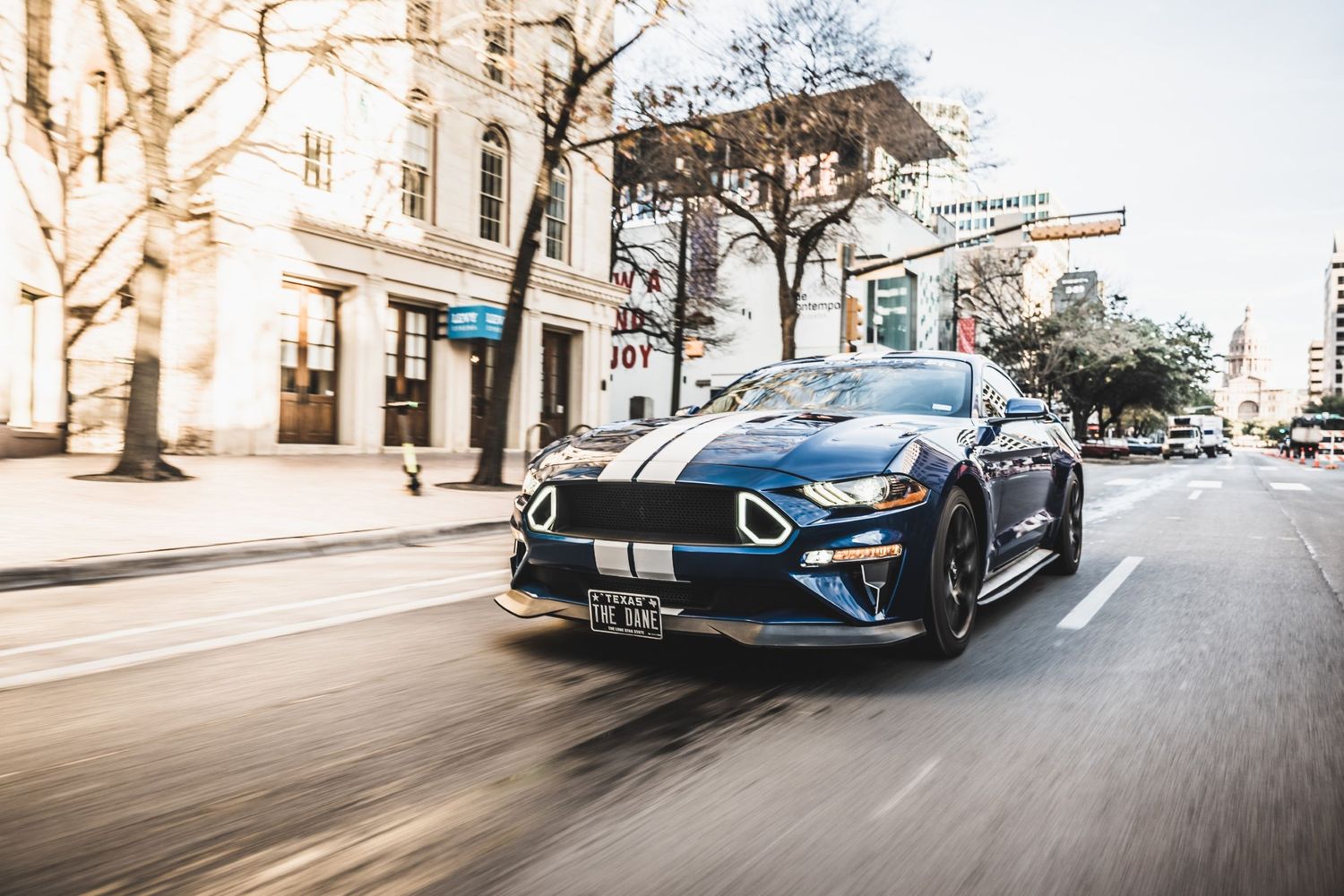 Blue Ford Mustang GT with racing stripes speeds through downtown streets.