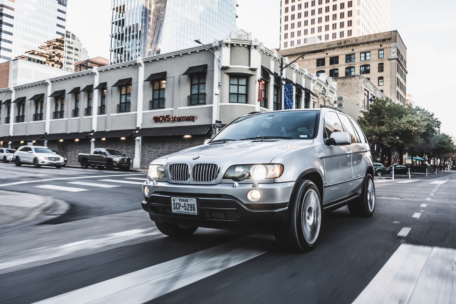 Silver BMW X5 SUV driving through downtown city streets with historic buildings visible.