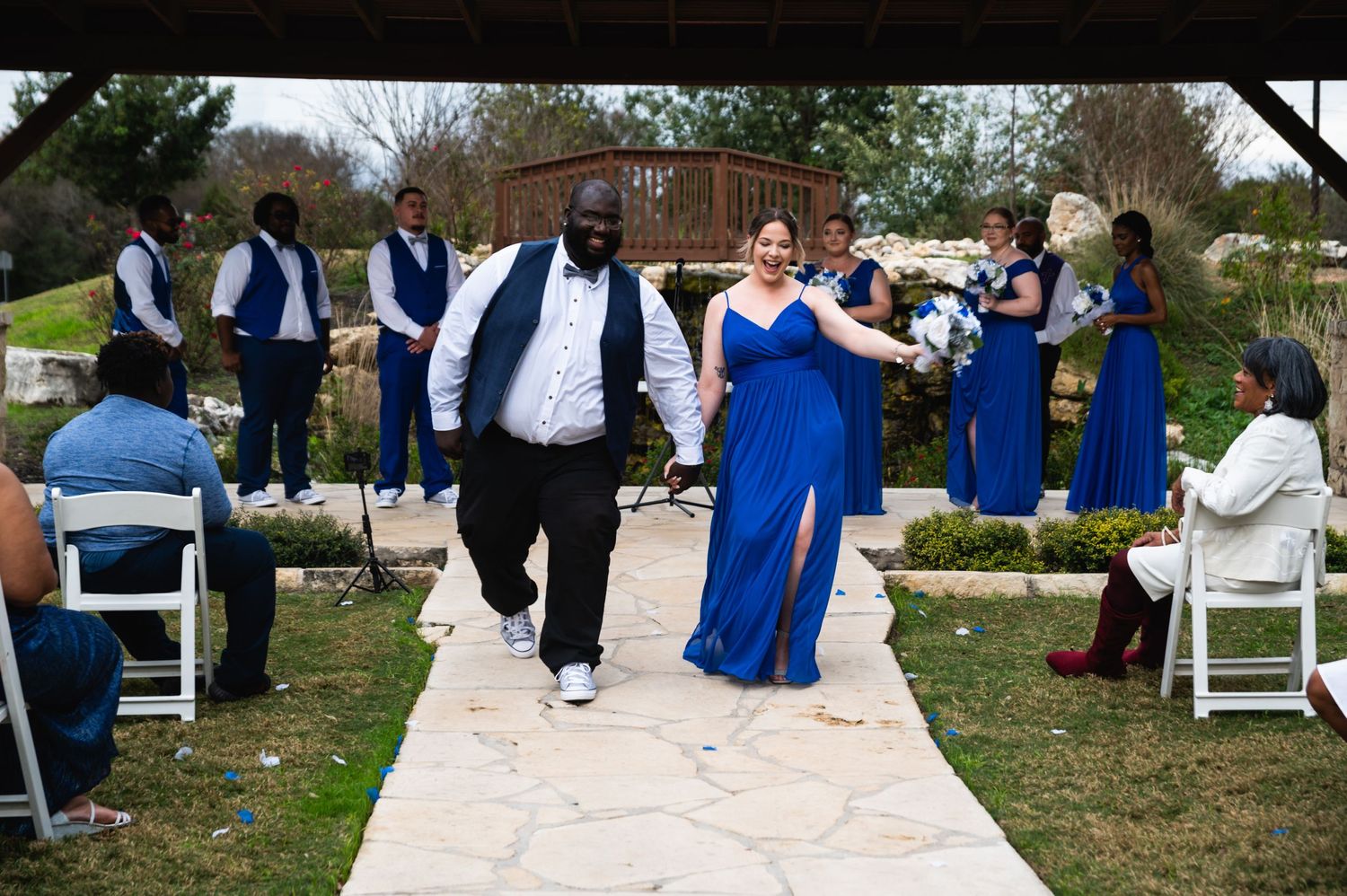 Wedding party members celebrate while walking down an outdoor aisle in coordinated royal blue and white outfits.