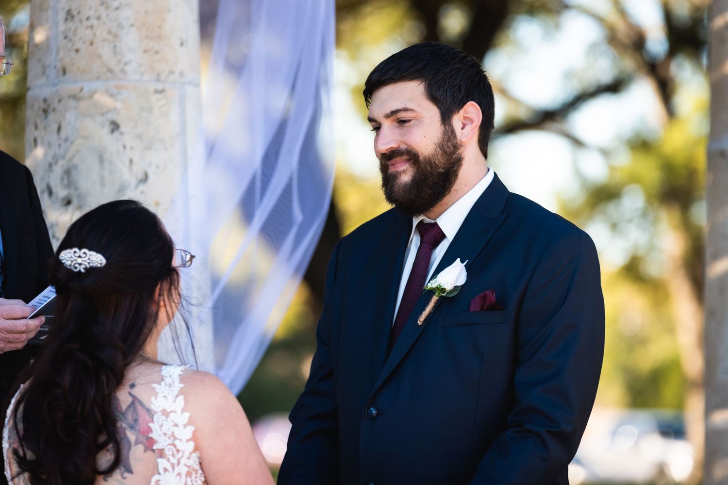 A wedding ceremony takes place outdoors with columns and drapery in the background.
