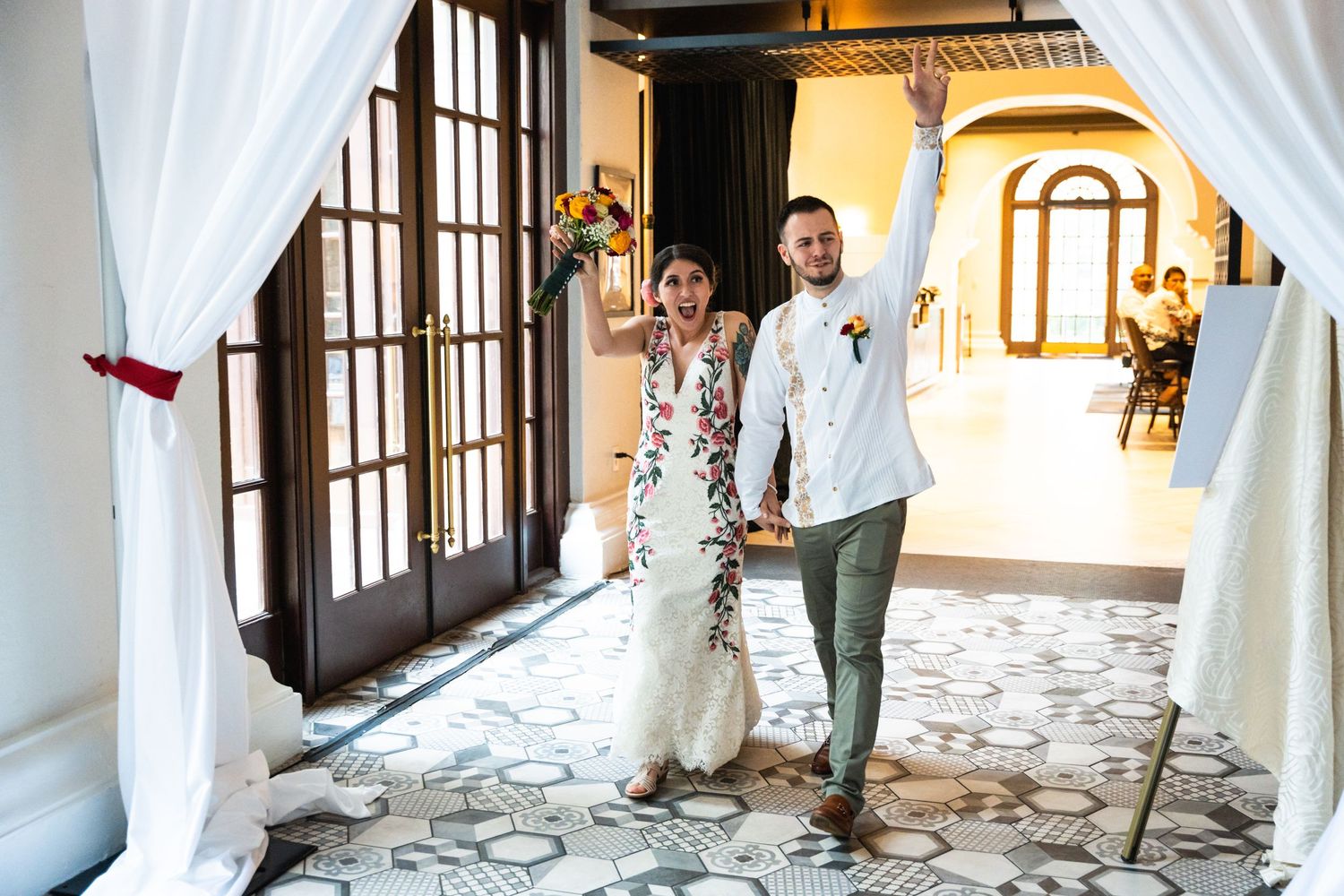 A couple walks through an elegant archway with white draping and stone columns.