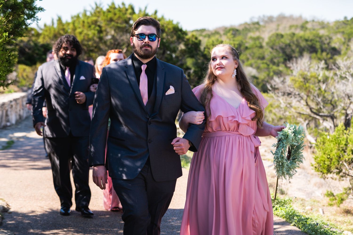Wedding party members walk along a sunny outdoor path wearing formal attire.