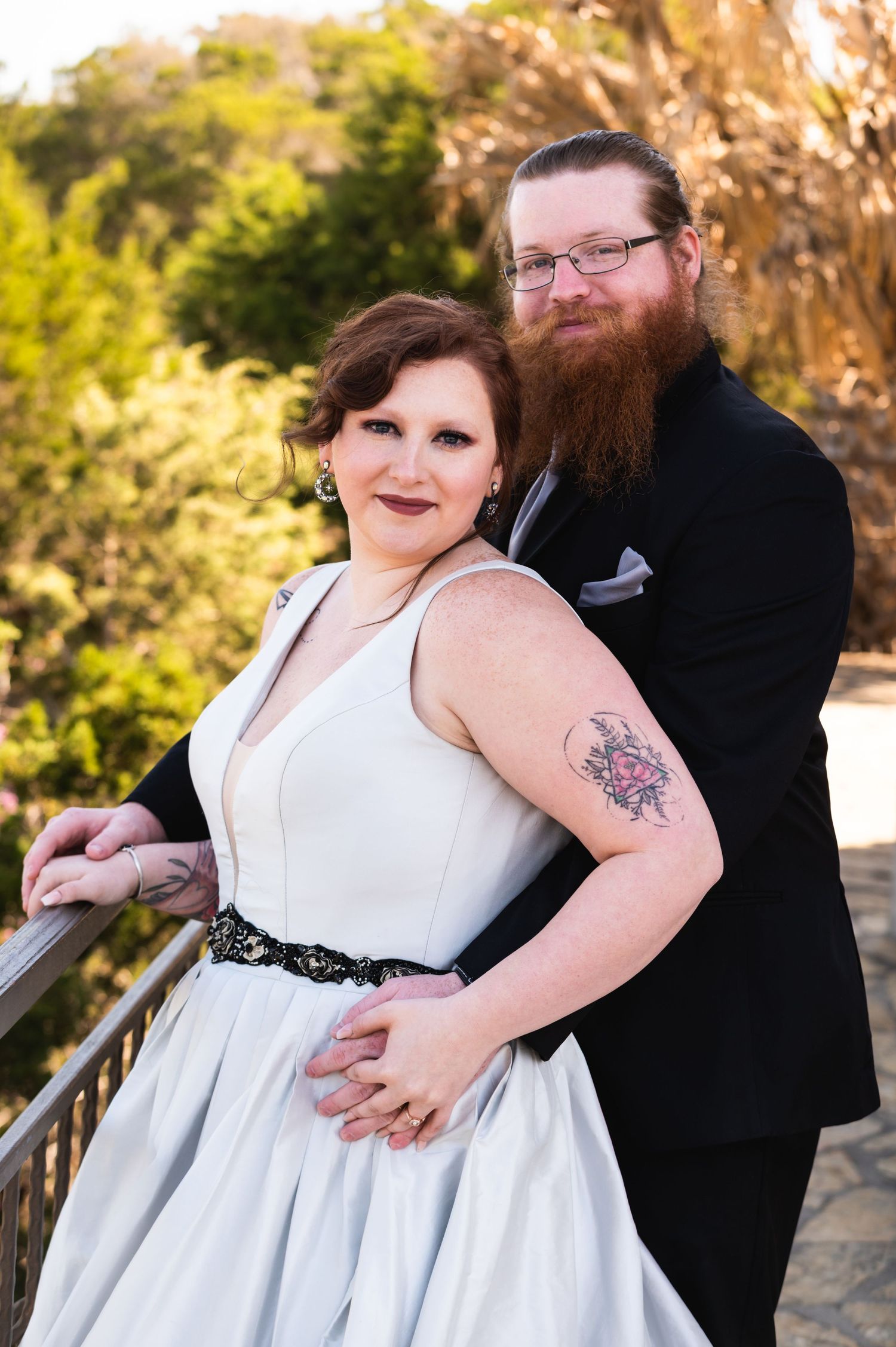 Newlyweds pose outdoors in formal attire against a sunny natural backdrop.