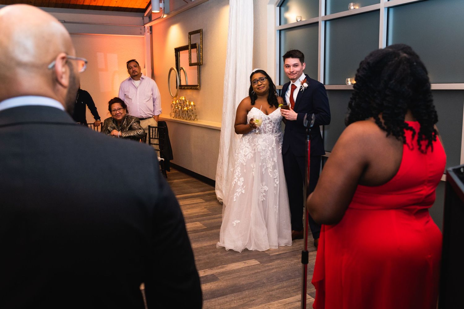 Wedding guests gather around the bride in a white lace gown during an indoor celebration.