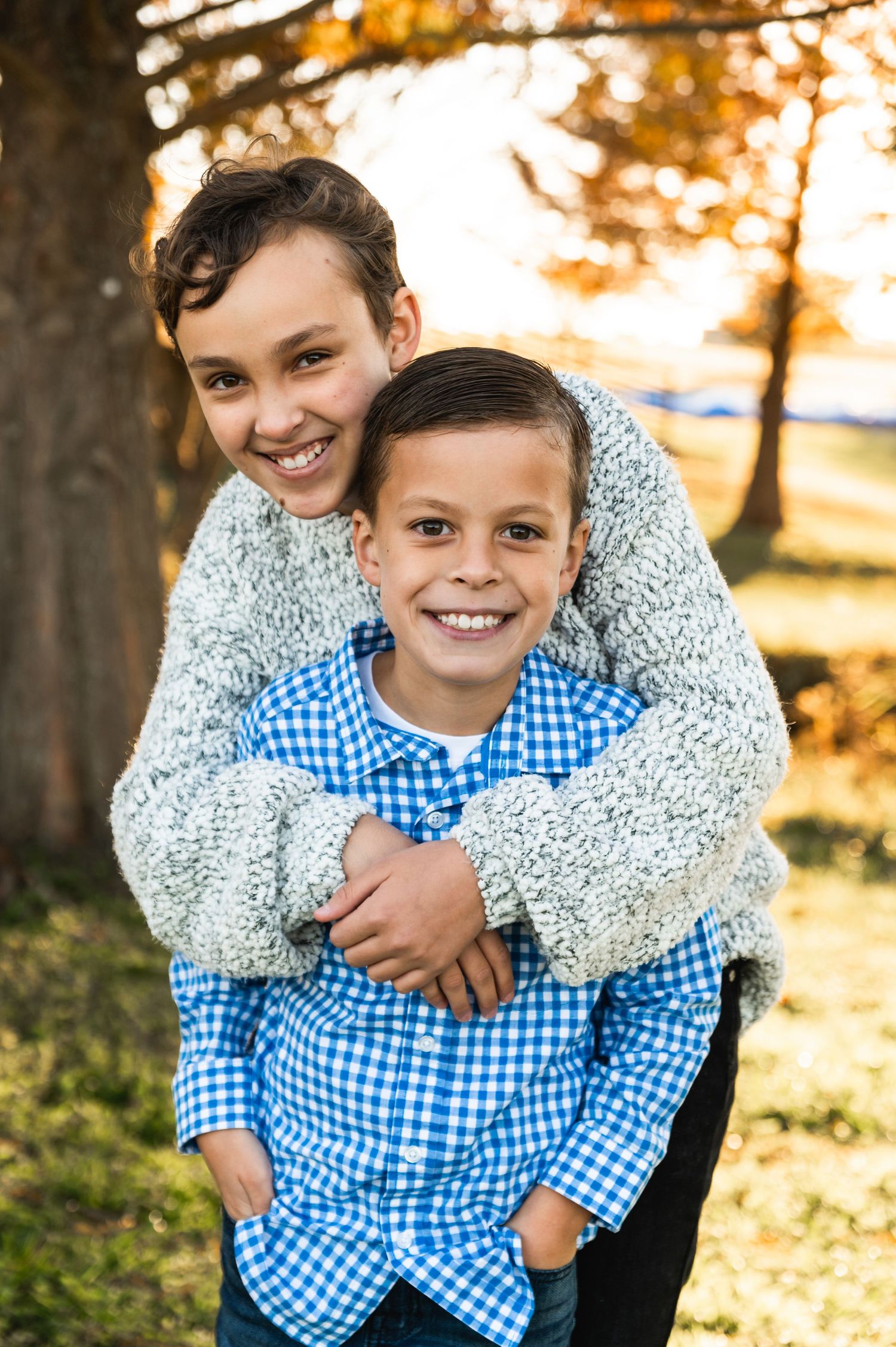Two people embrace while standing outdoors during golden hour with autumn trees in the background.