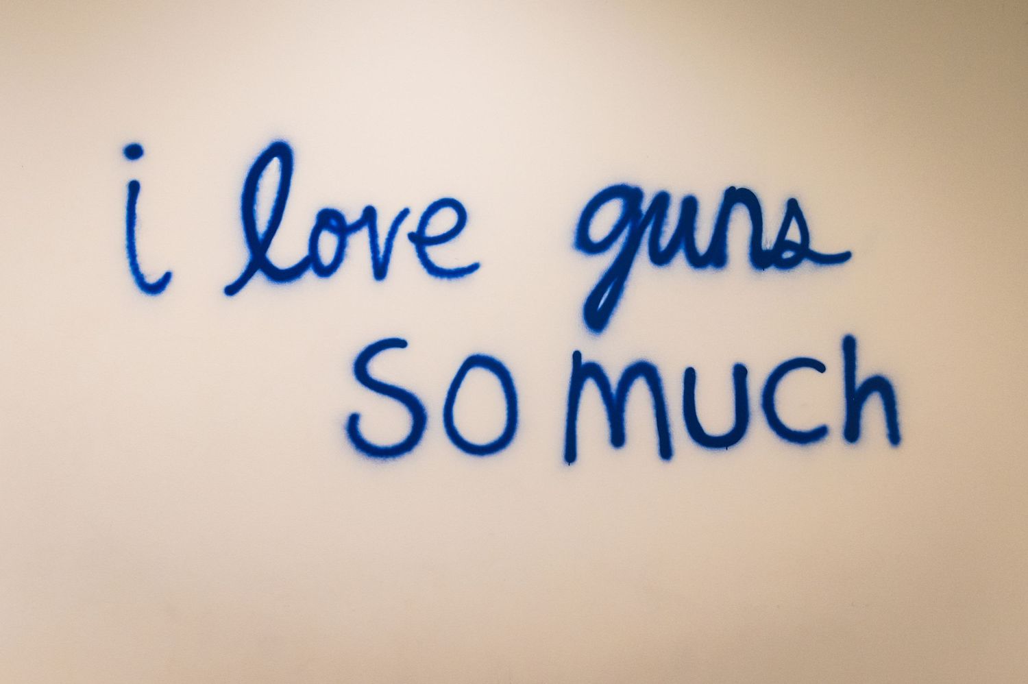 Blue handwritten text on a white wall reading 'i love guns so much'.