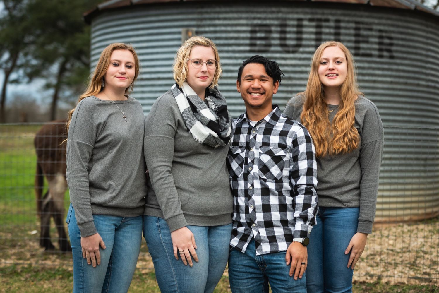 Four people in matching outfits pose together near metal grain silo in rural setting.