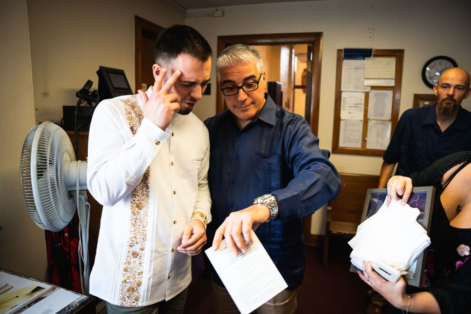 Two people examine documents together in an office setting with a fan visible in the background.